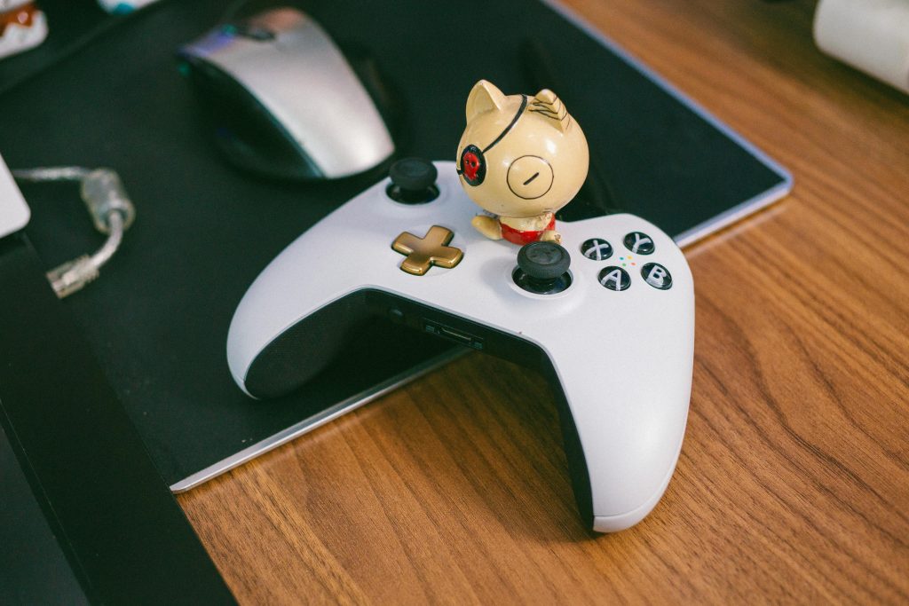 A charming cat figurine sits atop a gaming controller, beside a computer mouse, on a wooden desk.